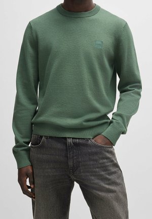 Homme portant un pull vert à manches longues avec un col rond et un petit patch logo carré sur la poitrine, et un jean foncé délavé.