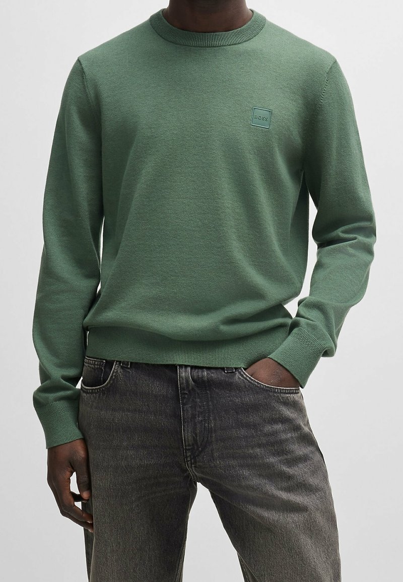 Homme portant un pull vert à manches longues avec un col rond et un petit patch logo carré sur la poitrine, et un jean foncé délavé.