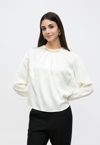 Hvid bluse med lange ærmer, rynket halsudskæring pyntet med guldbånd, glat tekstur og en let buet kant.