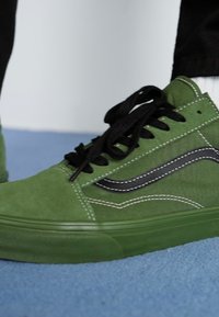 Zapatilla de ante verde con acentos negros y costuras blancas. Presenta un perfil bajo, puntera redonda y cordones negros. Superficie texturizada.