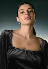 Top in satin nero con scollatura quadrata, corpetto arricciato e rifinitura argento decorata. La modella indossa orecchini a goccia blu e trasparenti.