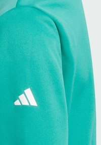 Tejido de sudadera turquesa con una textura suave. Presenta un logotipo blanco de Adidas en la manga izquierda. Vista acercada del área del hombro.