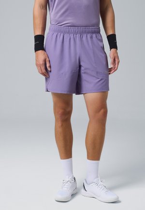 Uomo che indossa pantaloncini sportivi viola, scarpe da ginnastica bianche, calzini bianchi e polsini neri, in piedi su uno sfondo semplice.