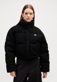 Svart, fluffy fleece pufferjakke med høy krage, framsideglidelås og quiltet design. Har en liten logo på venstre side.