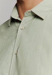 Lichtgroene linnen shirt met een puntkraag en een subtiel logo. Heeft zichtbare stiksels en ronde knopen in een contrasterende kleur.