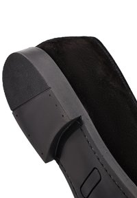 Chaussure noire en suède à enfiler avec une semelle en caoutchouc texturée, présentant un détail de couture subtil et un design incurvé au niveau du talon.