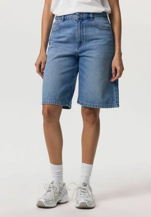 Kobieta nosząca niebieskie jeansowe spodenki o średniej długości, białe skarpetki i białe buty sportowe, stojąca na tle jednolitego jasnego tła.