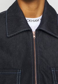 Mörk denimjacka med ett kopparfärgat blixtlås, blåa sömdetaljer och två framfickor med en klassisk krage.