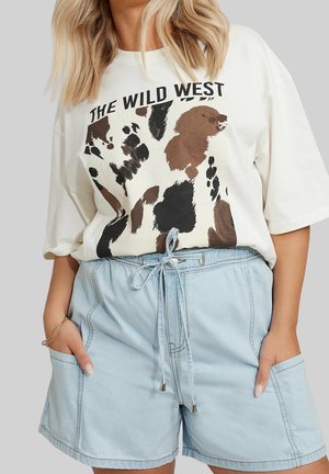 Kvinde iført en oversized hvid T-shirt med teksten "The Wild West" og ko-mønster, kombineret med lyseblå denim shorts med snøring.