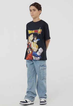 T-shirt nera a maniche corte con grafica di "Dragon Ball Super" che presenta personaggi colorati, abbinata a pantaloni cargo blu chiaro e sneakers.