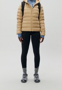 Veste beige matelassée avec capuche, leggings noirs, chaussettes grises et chaussures de randonnée grises/bleues. Sac à dos sur les épaules. Texture lisse, design fonctionnel.