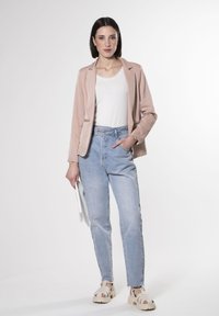 Hellrosa Blazer über einem weißen, spitzenbesetzten Top, kombiniert mit hoch taillierten blauen Jeans und beigen Sandalen. Hält eine weiße Clutch.