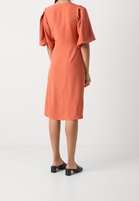 Robe couleur rouille avec des manches bouffantes, longueur genoux, texture lisse, taille cintrée, associée à des sandales noires à talons carrés.