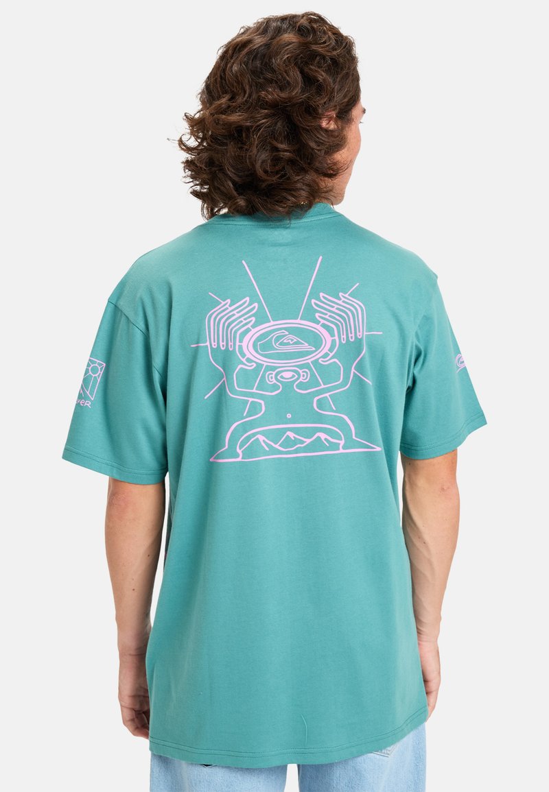 Mátově zelené bavlněné tričko s růžovým grafickým designem na zádech, znázorňující ruce a abstraktní tvary, s krátkými rukávy a volným střihem.