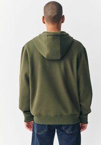 Person steht mit dem Rücken zur Kamera, trägt einen olivgrünen Hoodie und dunkle Jeans vor einem schlichten weißen Hintergrund.