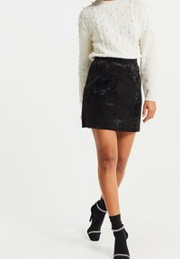 Vit tröja med texturerad kabelstickning och pärlprydnader, kombinerad med en svart sammet mini-kjol. Modellen har på sig svarta strumpor och klackskor.