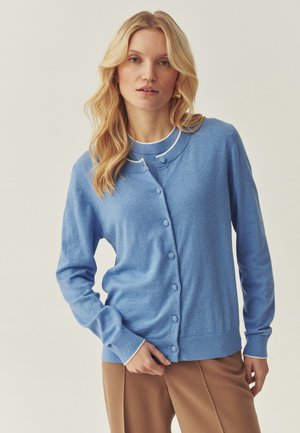 Cardigan en maille bleue avec un col rond, fermeture à boutons sur le devant, poignets et ourlet côtelés. Texture lisse, porté sur un haut blanc.