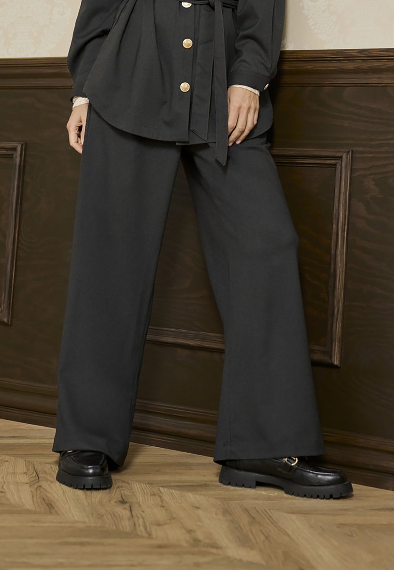 Weite schwarze Hosen mit glatter Textur, kombiniert mit klobigen schwarzen Schuhen mit Riemen und strukturiertem Sohlen, vor einem Hintergrund aus Holzpaneelen.