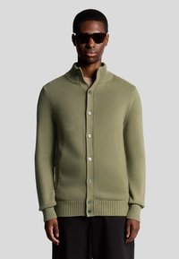 Cardigan en maille vert kaki avec un grand col, cinq boutons, des poignets et un ourlet côtelés, porté sur une chemise de couleur claire.