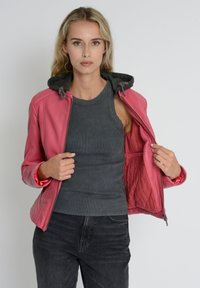 Lyserød læderjakke med grå hætte, lynlås-lukning og quiltet inderfor. Modellen bærer en grå tanktop og mørke jeans, mens hun holder jakken åben.