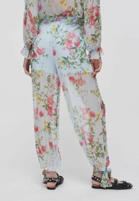 Pantalon transparent à fleurs roses et jaunes, noué aux chevilles, porté avec des sandales noires cloutées, vu de dos sur un fond uni.