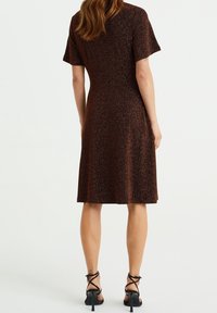 Kvinna iklädd en knälång, kortärmad, brun strukturerad klänning med svarta remmar och högklackade sandaler, stående med ryggen vänd.