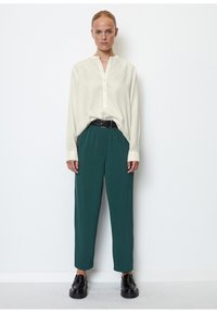 Langärmlige, off-white Bluse mit Henley-Kragen und Knopfblende, kombiniert mit tealfarbenen, engen Hosen und schwarzen Plateauschuhen.