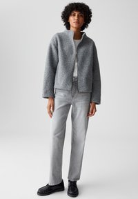 Giacca in pile grigia con colletto alto, zip davanti e maniche ampie, abbinata a jeans dritti grigio chiaro e scarpe nere.