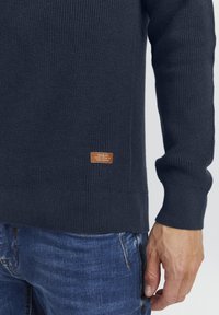 Pull en tricot bleu marine avec un design texturé, des poignets et un ourlet côtelés. Comprend une petite étiquette marron avec du texte près du coin inférieur gauche.