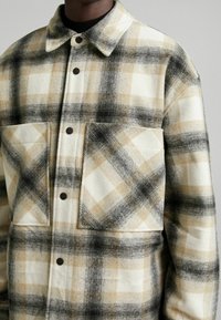 Detalle en primer plano de una camisa de cuadros en beige, negro y crema, con dos grandes bolsillos frontales y botones de presión, puesta por una persona.