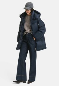 Marineblauwe pufferjack met capuchon, geribde grijze trui, wijde donkere denim broek en een grijze pet. Kenmerken zijn ritsen en subtiele stikdetails.