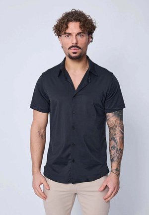 Homme aux cheveux bouclés et à la barbe portant une chemise noire à manches courtes boutonnée et un pantalon beige, montrant une manche tatouée sur son bras droit.