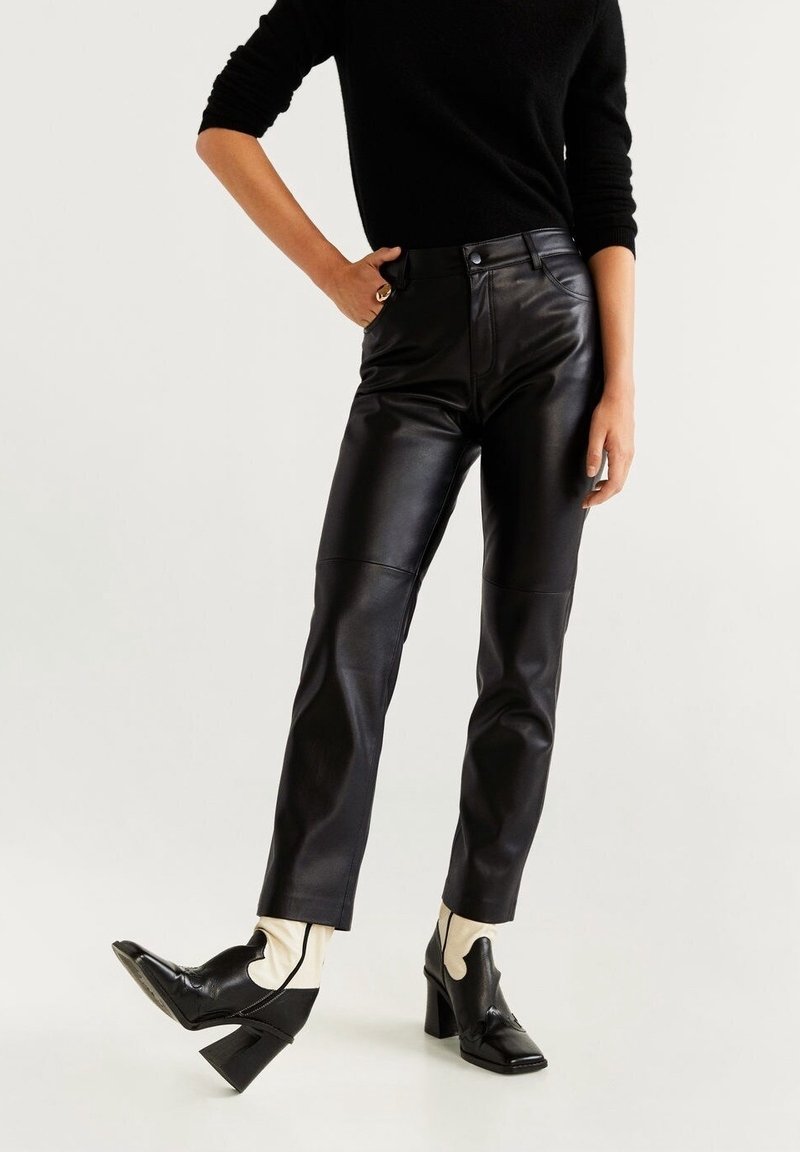 Personne portant un pantalon en cuir noir, des bottines à talons noires et blanches, et un haut noir à manches longues, posant avec une main sur la hanche.