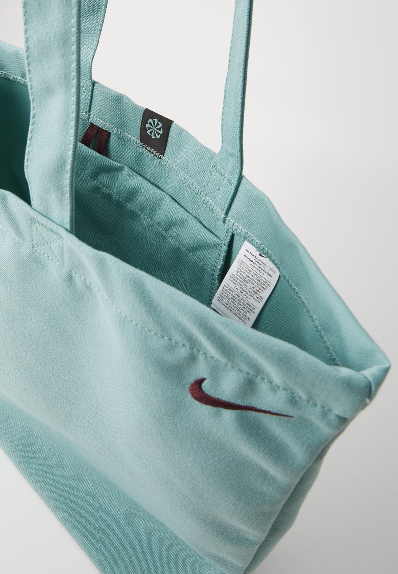 Bolsa tote en color verde azulado claro con dos asas, que presenta un logo de Nike en color marrón y una etiqueta negra en el interior. Hecha de una tela suave con una textura lisa.