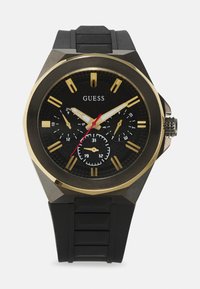 Montre avec bracelet en silicone noir et boîtier octogonal noir et or. Elle présente trois sous-cadrans, de grands index horaires dorés et une aiguille des secondes rouge.