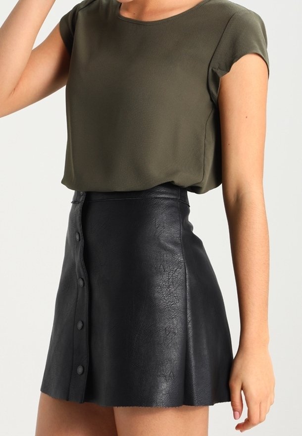 Olijfgroene blouse met korte mouwen, gecombineerd met een zwarte neplederen minirok met een sluiting met knopen aan de voorkant en een gladde, gestructureerde afwerking.
