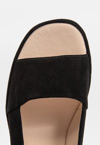 Sandale en suede noire avec une large bride supérieure et une semelle intérieure lisse beige. Elle présente un design à bout carré et des détails de coutures minimalistes.