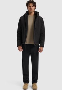 Schwarze isolierte Jacke mit Kapuze, beiger Pullover, schwarze Hose und braune Schuhe. Glatte Stoffqualität mit strukturierter Passform.