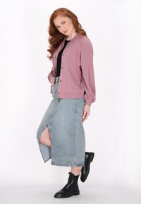 Giubbotto bomber rosa chiaro con bottoni a pressione, abbinato a una gonna di jeans con spacco alto. Il modello indossa stivaletti neri alla caviglia. Tessuto dalla consistenza liscia.