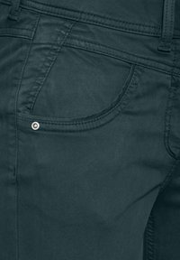 Tmavě zelená denimová tkanina s hladkým povrchem. Obsahuje zakřivenou kapsu se stříbrným nýtem a švy s detailním prošíváním.