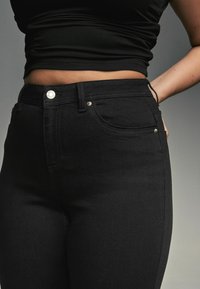 Čierne džínsy z denimu so slim fit strihom, predným vreckom, jemným prešívaním a strieborným gombíkom na páse. Hladká textúra.