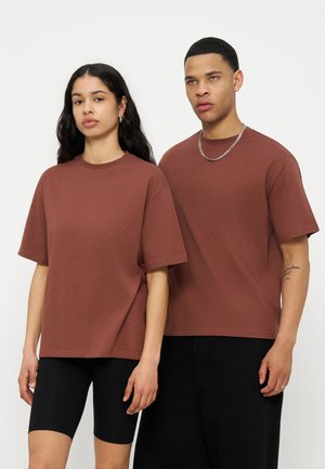 Fiatal nő és férfi, egyforma barna, túlméretezett pólókat, fekete rövidnadrágot és nadrágot viselve, egymás mellett állnak fehér háttér előtt.