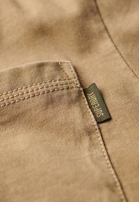 Beige cotton fabric with a visible pocket, featuring a green "Superdry" label. The texture is smooth, with fine stitching details.