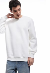 Sudadera blanca de cuello redondo hecha de una tela suave, con mangas largas, un corte relajado y puños y dobladillo de canalé. Combinada con unos pantalones vaqueros claros.