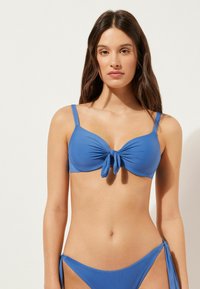 Blå bikini med en knutdetalj i mitten, justerbara spännen och slät tyg. Designen inkluderar en klassisk trekantsform.