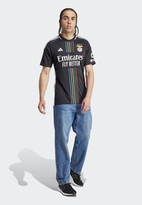 Maglietta nera a maniche corte Adidas con strisce verticali colorate, con la scritta "Emirates Fly Better" e un emblema a tre stelle. Indossata con jeans blu.