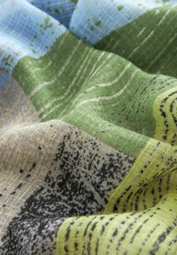 Tissu texturé avec des blocs verts, bleus, jaunes et beige, présentant des motifs tachetés et rayés noirs, montré en gros plan avec des plis.