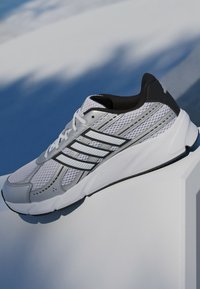 Hvid atletiksko med mesh-overdel og sorte detaljer. Har tre fremtrædende hvide striber og et tekstureret ydersål-design. Letvægtsdesign.