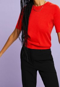 Personne portant un pull rouge vif à manches courtes, un pantalon noir, un collier chaîne doré et de longs cheveux noirs tressés sur fond violet.