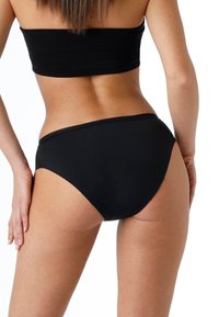 Donna che indossa mutandine nere a vita alta e un top nero senza spalline abbinato, vista da dietro su sfondo bianco.